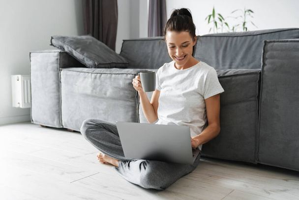 Smiling woman drinking coffee and using laptop while sitting on floor at home - Φωτογραφία, εικόνα