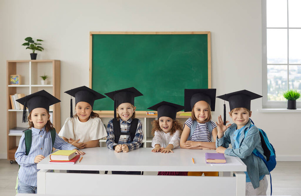 Group of happy school children in graduation caps standing by table with books in classroom - Foto, imagen