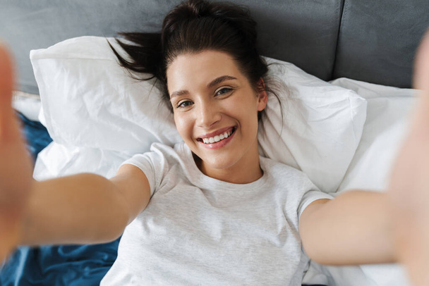 Brunette woman smiling and taking selfie photo while lying on bed at home - Fotó, kép