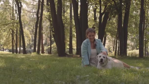 Lange Aufnahme einer jungen kaukasischen Frau im hellblauen Blazer, die auf dem Rasen im sonnigen Park sitzt, ihren lieblichen Hund streichelt und dann davonläuft - Filmmaterial, Video