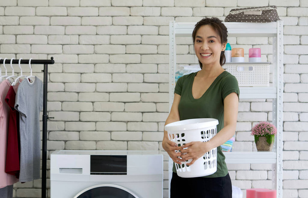 Asiática ama de casa sosteniendo cesta de plástico delante de la lavadora. Ambiente de trabajo en la lavandería. - Foto, Imagen