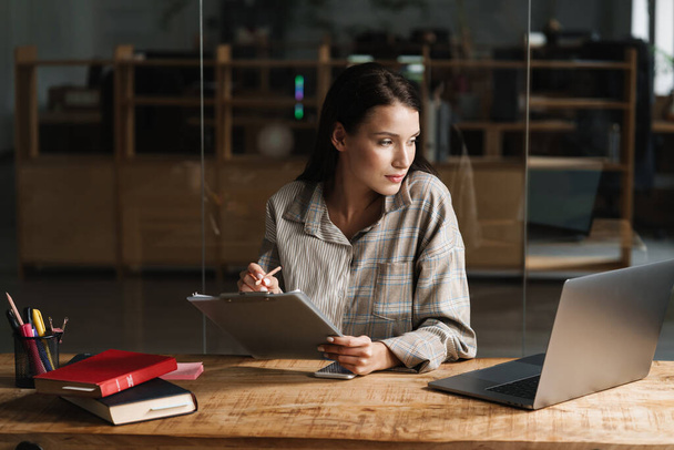 Young brunette woman writing down notes while working with laptop in office - Foto, immagini