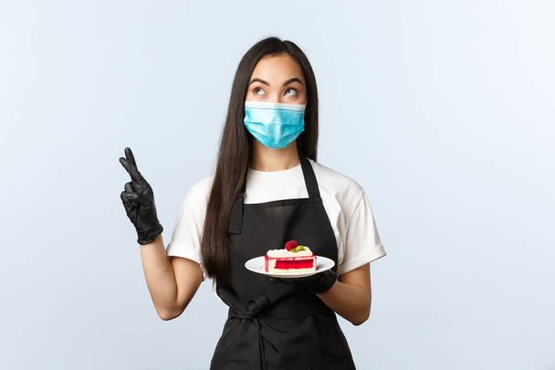 Covid-19, social distancing, small coffee shop business and preventing virus concept. Hopeful cute asian waitress, barista or cafe owner in medical mask cross fingers, hold plate with cake - Photo, Image