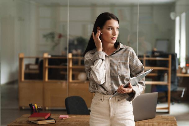 Mid aged casual white brunette woman standing in office with clipboard wearing earphone - Photo, Image