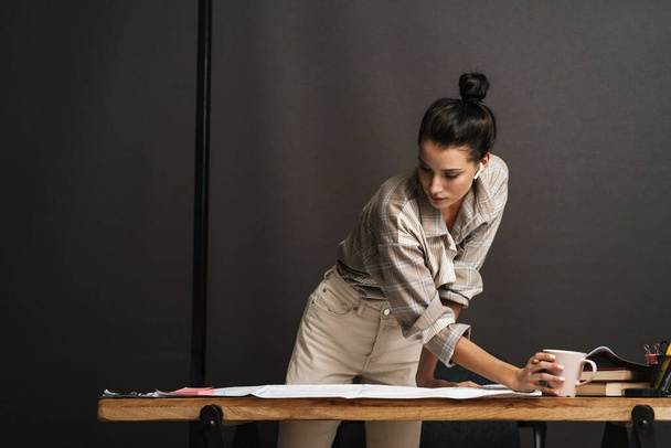 Young brunette woman drinking coffee while working with drawings in office - Foto, imagen