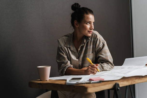 Young brunette woman working with drawings while sitting at desk in office - Fotoğraf, Görsel