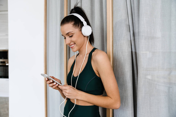 Smiling young white woman in sportswear in headphones listening to music holding mobile phone standing in the living room - Фото, изображение