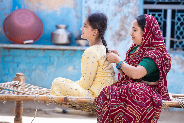 農村部のインド人の母親は、村の家の伝統的なベッドに座っている間、彼女の若い愛らしい娘の髪を編みます。可愛いです就学前の子供とともに過ごします,赤いサリーの女性と黄色のドレスの女の子 - 写真・画像