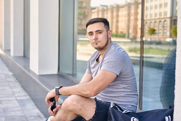 athletischer junger Kaukasier mit dunklen Haaren sitzt vor einem Gebäude und stützt seine Ellbogen auf die Beine - Foto, Bild