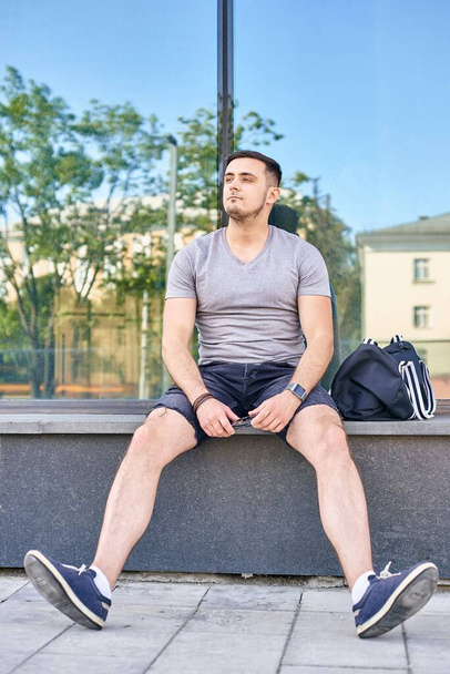αθλητικός νεαρός καυκάσιος άνδρας με σκούρα μαλλιά κάθεται δίπλα στο κτίριο και κουρασμένος μετά την προπόνηση - Φωτογραφία, εικόνα