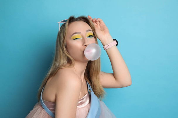 Fashionable young woman with bright makeup blowing bubblegum on light blue background - Fotó, kép