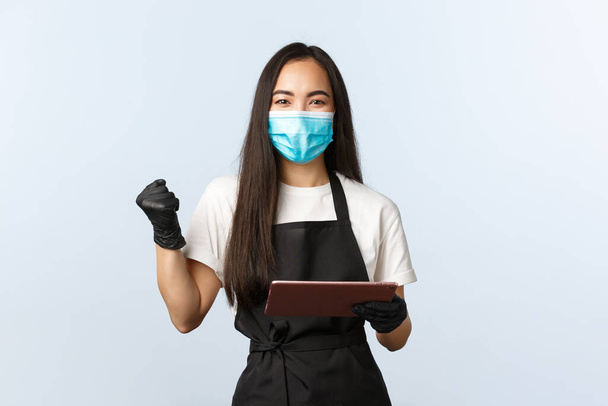 Covid-19, online orders, small coffee shop business and preventing virus concept. Satisfied rejoicing asian female barista, cafe worker fist pump as using digital tablet, wear medical mask - Valokuva, kuva