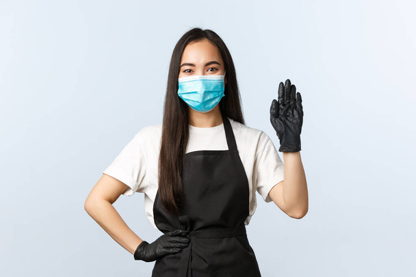 Covid-19-Pandemie, soziale Distanzierung, Kleinunternehmen und Virusprävention. Freundliche Barista in medizinischer Maske und Handschuhen grüßt Kunden, begrüßt Verbraucher und bestellt im Café - Foto, Bild