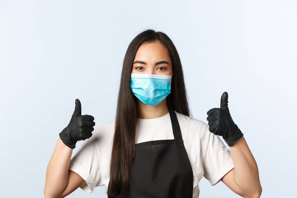 pandemia de Covid-19, cafetería, pequeña empresa y prevención del concepto de virus. Asiática mujer dueña de café en máscara médica y guantes, barista show thumb-up, invitar a vistitors - Foto, Imagen