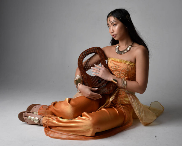 Full length portrait of pretty young asian woman wearing golden Arabian robes like a genie, holding a small musical harp, isolated on studio background. - 写真・画像