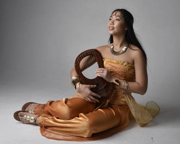 Ganztägiges Porträt einer hübschen jungen Asiatin, die goldene arabische Gewänder trägt wie ein Genie, eine kleine musikalische Harfe in der Hand, isoliert auf Studiohintergrund. - Foto, Bild