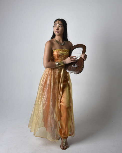 Full length portrait of pretty young asian woman wearing golden Arabian robes like a genie, holding a small musical harp, isolated on studio background. - Foto, afbeelding
