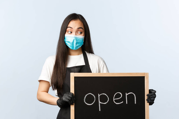 Small business, covid-19 pandemic, preventing virus and employees concept. Smiling cute asian female employee, cafe or store worker holding open sign, invite visitors - Photo, image