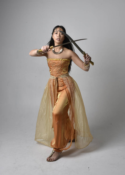 Ritratto a figura intera di bella giovane donna asiatica vestita con abiti arabi dorati come un genio, con in mano un'arma a spada, isolata sullo sfondo dello studio. - Foto, immagini