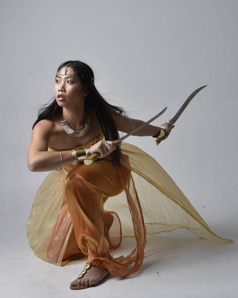 Full length portrait of pretty young asian woman wearing golden Arabian robes like a genie, holding a sword weapon, isolated on studio background. - 写真・画像