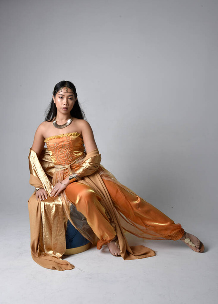 Full length portrait of pretty young asian woman wearing golden Arabian robes like a genie, seated pose, isolated on studio background. - Fotografie, Obrázek