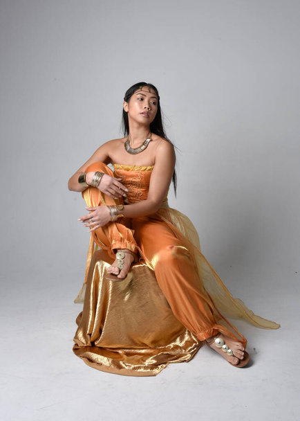 Retrato de larga duración de una hermosa joven asiática vestida con túnicas árabes doradas como un genio, pose sentada, aislada en el fondo del estudio. - Foto, Imagen