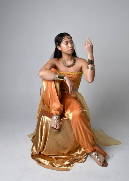 Full length portrait of pretty young asian woman wearing golden Arabian robes like a genie, seated pose, isolated on studio background. - Photo, image