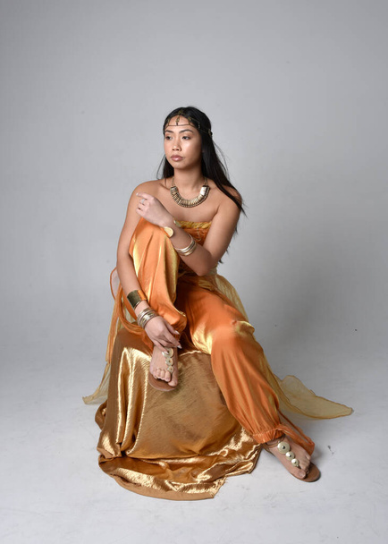 Ολο το μήκος πορτραίτο μιας όμορφης νεαρής Ασιάτισσας που φορούσε χρυσές αραβικές ρόμπες σαν τζίνι, καθισμένη πόζα, απομονωμένη στο φόντο του στούντιο. - Φωτογραφία, εικόνα
