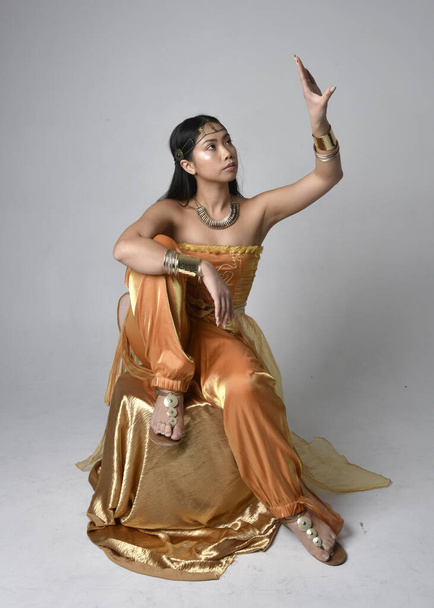 Teljes hossz portré csinos fiatal ázsiai nő visel arany arab köntös, mint egy dzsinn, ülő póz, elszigetelt stúdió háttér. - Fotó, kép