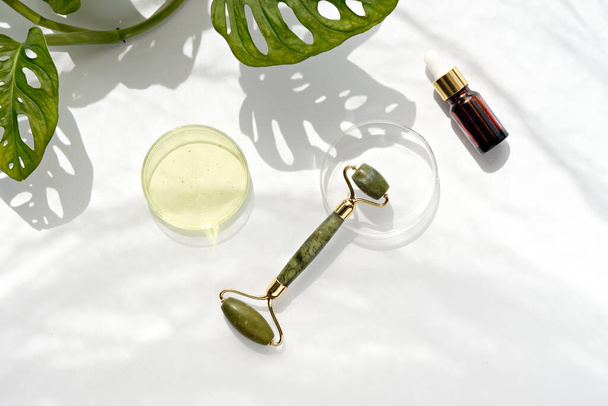 Moisturizer, green jade face roller with exotic leaves. Monstera Adansonii leaves. Sunshine, long shadows. Minimal flat lay on off white background. Facial massage with self made cosmetics. - Φωτογραφία, εικόνα