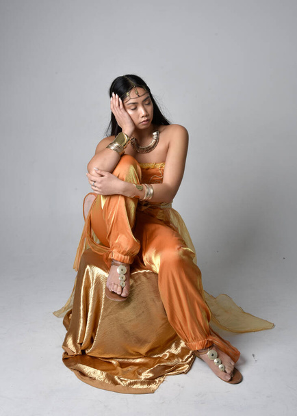 Portrait pleine longueur de jolie jeune femme asiatique portant des robes arabes dorées comme un génie, pose assise, isolée sur fond de studio. - Photo, image