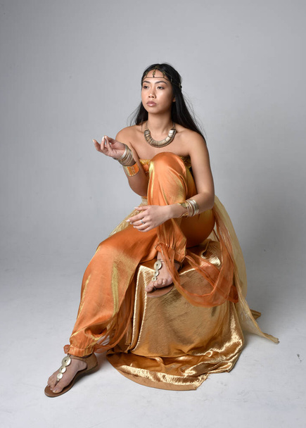 Full length portrait of pretty young asian woman wearing golden Arabian robes like a genie, seated pose, isolated on studio background. - Фото, зображення