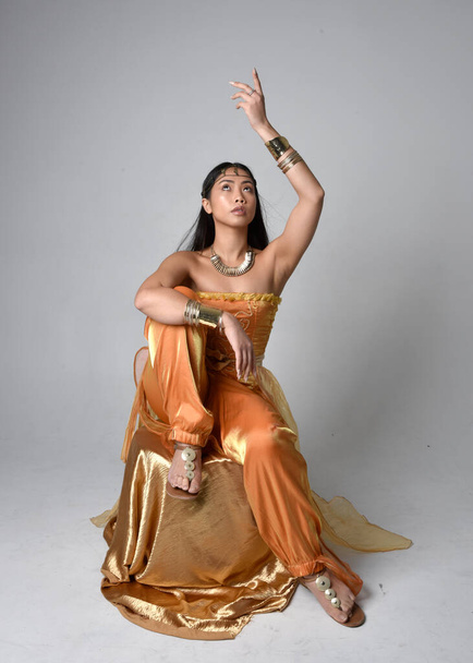 Retrato de larga duración de una hermosa joven asiática vestida con túnicas árabes doradas como un genio, pose sentada, aislada en el fondo del estudio. - Foto, imagen