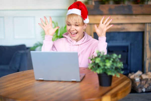 Χαρούμενη ενθουσιασμένη νεαρή γυναίκα με καπέλο Αϊ Βασίλη μπροστά στην οθόνη του φορητού υπολογιστή. Online συνάντηση με φίλους ή οικογένεια την παραμονή των Χριστουγέννων ή της Πρωτοχρονιάς. - Φωτογραφία, εικόνα