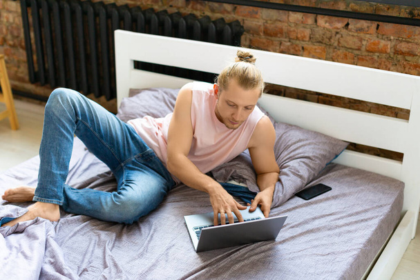 Un joven con un portátil en la cama. Concepto de tecnologías modernas en la vida cotidiana y en el trabajo remoto. - Foto, Imagen