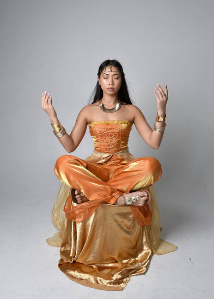 Ganzes Porträt einer hübschen jungen asiatischen Frau, die goldene arabische Roben trägt wie ein Genie, sitzend, isoliert auf Studiohintergrund. - Foto, Bild