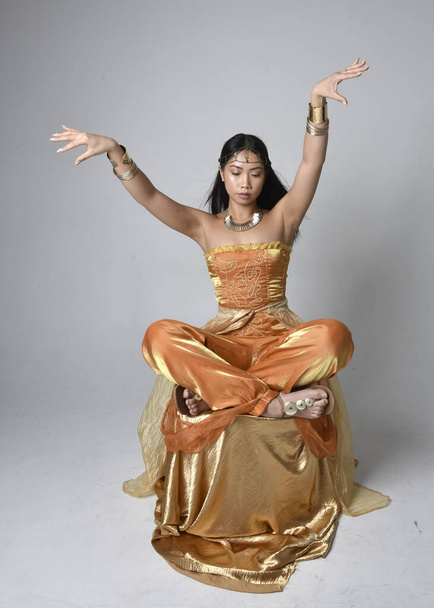 Портрет молодої азіатки, одягненої в золотий арабський одяг, як джин, сидячої пози, ізольованої на задньому плані студії.. - Фото, зображення