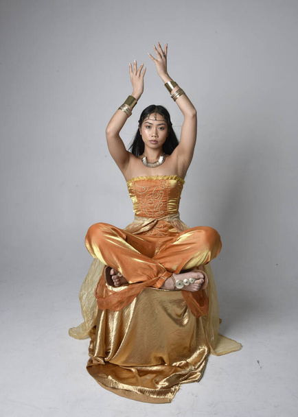 Full length portrait of pretty young asian woman wearing golden Arabian robes like a genie, seated pose, isolated on studio background. - Fotoğraf, Görsel
