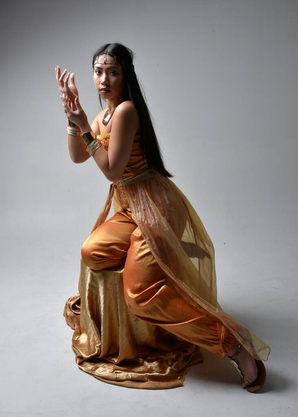Retrato de larga duración de una hermosa joven asiática vestida con túnicas árabes doradas como un genio, pose sentada, aislada en el fondo del estudio. - Foto, Imagen