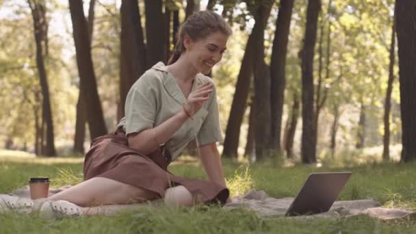 Pełne ujęcie młodej białej kobiety siedzącej na kocu na trawniku w słonecznym parku, dzwoniącej przez komputer przenośny - Materiał filmowy, wideo