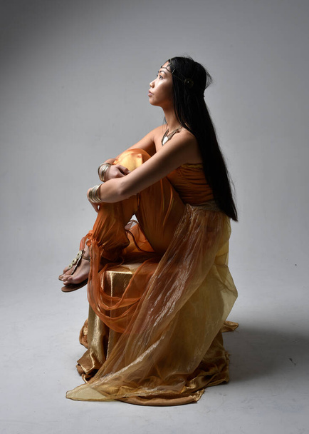 Portrait pleine longueur de jolie jeune femme asiatique portant des robes arabes dorées comme un génie, pose assise, isolée sur fond de studio. - Photo, image