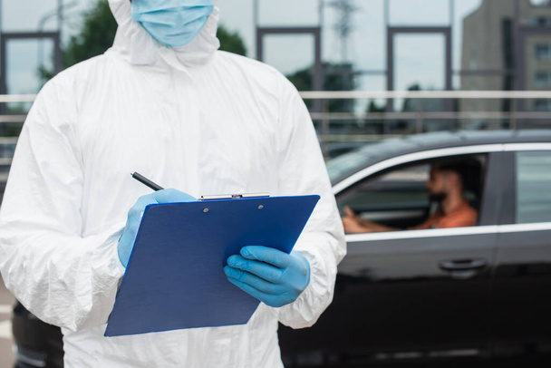 Travailleur médical en costume Hazmat écriture sur presse-papiers près de l'homme flou dans la voiture  - Photo, image