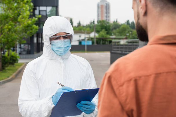 Trabajador médico con traje de materiales peligrosos escribiendo en el portapapeles cerca de un hombre borroso al aire libre  - Foto, Imagen