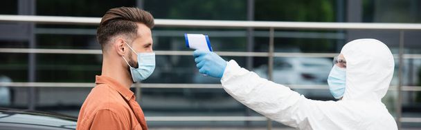 Trabajador médico sosteniendo pirómetro cerca del conductor en máscara médica al aire libre, pancarta  - Foto, imagen