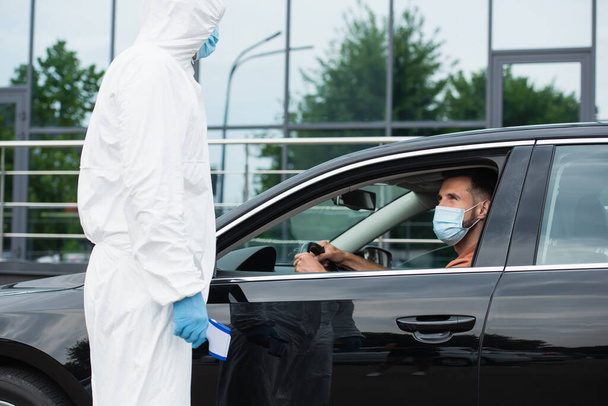 Medical worker in protective suit holding pyrometer near driver in car  - Fotoğraf, Görsel