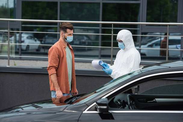 Медичний працівник у костюмі небезпеки, що пише на кишені біля людини в масці та авто на відкритому повітрі
  - Фото, зображення