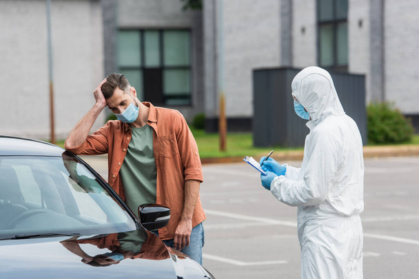 Sad driver standing near medical worker in hazmat suit writing on clipboard  - Photo, Image
