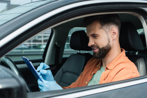 Motorista sorridente em luvas de látex escrevendo na área de transferência no carro  - Foto, Imagem