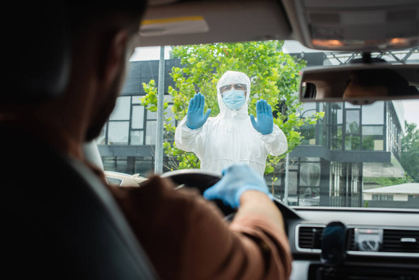 Trabajador médico en traje protector mostrando parada en conductor borroso en auto  - Foto, imagen