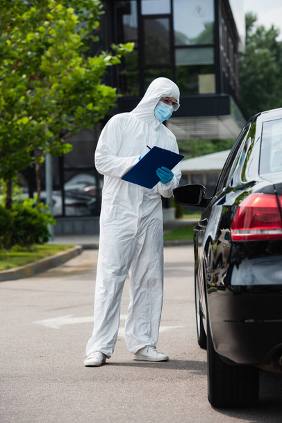 Tehlikeli madde giysisi giymiş bir tıp çalışanı dışarıda arabanın yanında panoya yazı yazıyor.  - Fotoğraf, Görsel